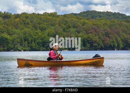 Senior man with hat, fishing from a small canoe on Limekiln Lake in Old Forge, New York. He's reeling in a cast. Stock Photo