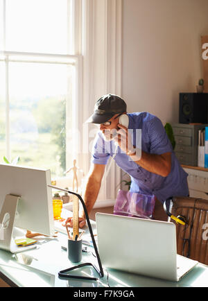 Man talking on telephone and using computer at desk in sunny home office Stock Photo