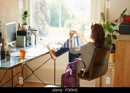 Pensive woman looking through window with feet up on desk in sunny home office Stock Photo