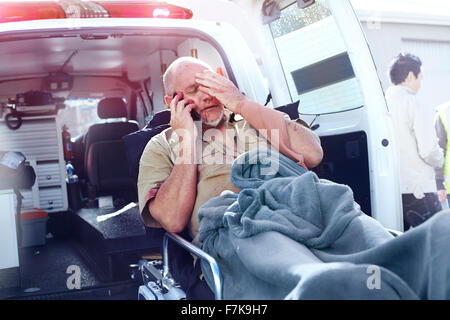 Distressed man on stretcher talking on cell phone behind ambulance Stock Photo