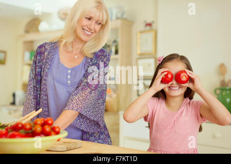 Portrait playful granddaughter covering eyes with tomatoes in kitchen Stock Photo