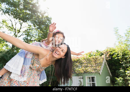 Carefree mother piggybacking daughter with arms outstretched in backyard Stock Photo