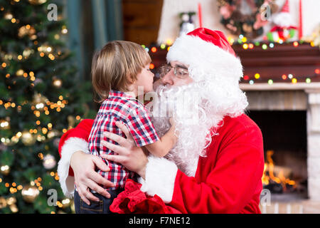 Child toddler boy kissing Santa Claus sitting near Christmas tree Stock Photo