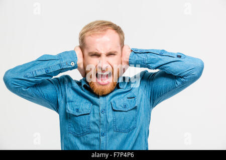 Mad hysterical young man with beard closed ears by hands and screaming isolated over white background Stock Photo