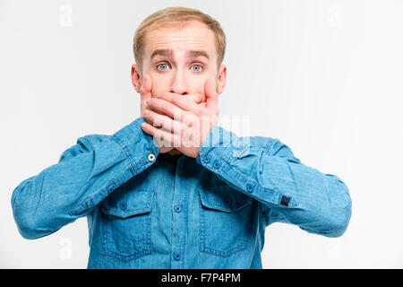 Shocked dazed young blond man closed his mouth by hands over white background Stock Photo