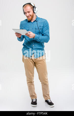 Positive attractive young male with beard in jeans shirt and beige pants listening to music from tablet using headphones over wh Stock Photo