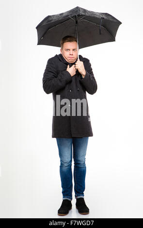 Amusing young man in black coat and jeans standing and feeling cold under umbrella over white background Stock Photo