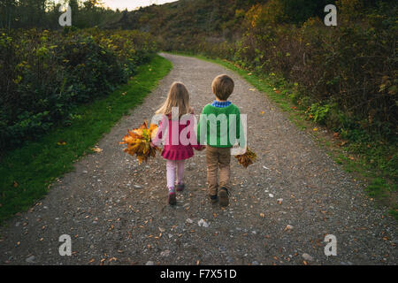 Boy and girl walking down path holding hands, carrying autumn leaves Stock Photo