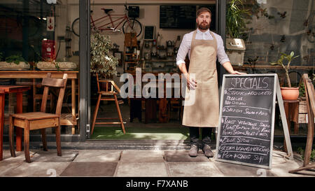 Portrait of young man standing at the entrance of his coffee shop with a black board. Barista standing in the doorway of a resta Stock Photo