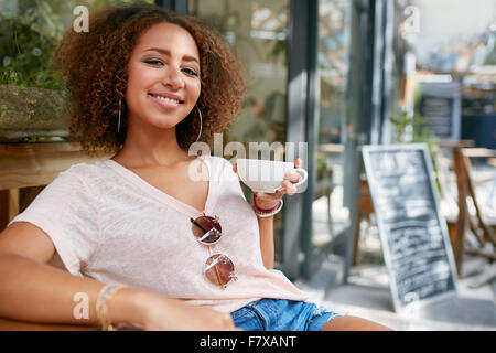 Portrait of young woman drinking coffee. African woman sitting at cafe holding a cup of coffee, looking at camera smiling. Stock Photo