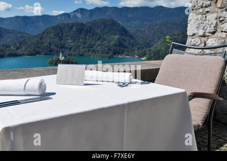 Served table with empty plate outdoor. View over lake Bled, island with church and Alps mountains in the background. Slovenia. B Stock Photo