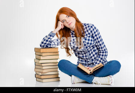 Thoghtful beautiful young curly redhead girl sitting with legs crossed and leaning on stack of the books over white background Stock Photo
