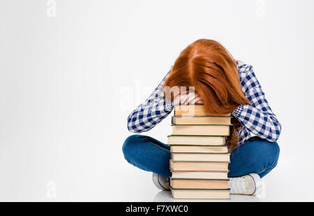 Tired exhausted young woman with red hair put head on stack of books and sleeping over white background Stock Photo