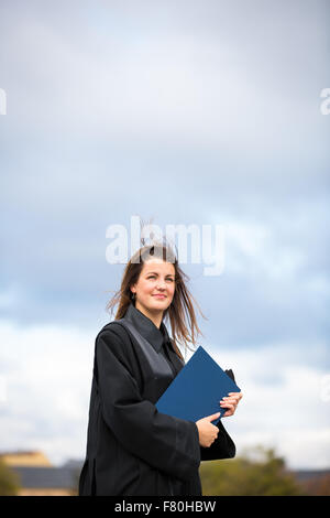 Pretty, young woman celebrating joyfully her graduation - spreading wide her arms, holding her diploma, savouring her success (c Stock Photo