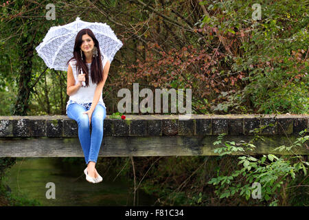 Beautiful brunnette sitting on bridge holding white lace umbrella. Stock Photo