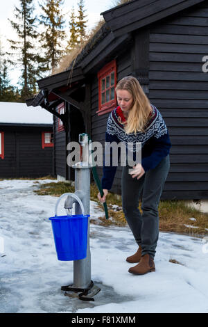 Pretty girl pumping a hand pump to fill a bucket from a well outside a cabin in the forest North of Oslo Stock Photo