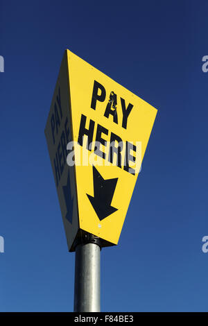 A sign points people to 'Pay Here' at a car park in Lewes, England. The yellow sign is contrasted against the blue sky.