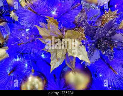 Close up of gold colored poinsettia hanging in white Christmas tree with several other ornaments on blue background. Stock Photo