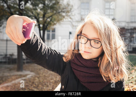 Beautiful Caucasian blond teenage girl taking photo on her smartphone in autumnal park, vintage style tonal correction photo Stock Photo