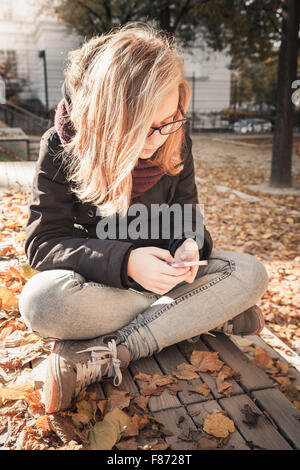 Cute Caucasian blond teenage girl in jeans and black jacket sitting on wooden park bench and using smartphone, outdoor autumn Stock Photo