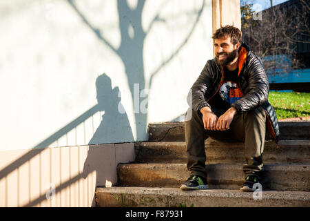 brown bearded young boy sitting on stairs street bestido with feathery gray and orange , a black T-shirt, green pants and sneake Stock Photo