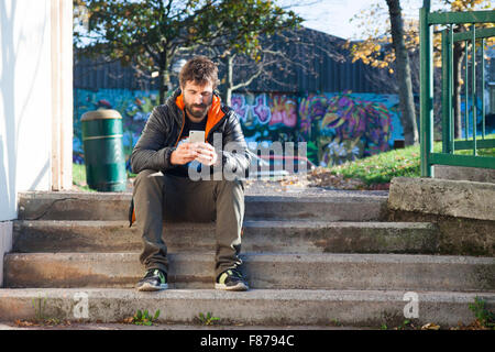 brown bearded young boy sitting on stairs street using his smartphone , wearing a gray and orange hack , a black T-shirt, green Stock Photo