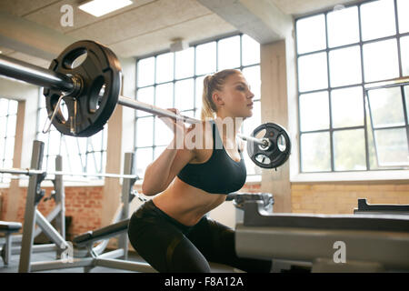 Female exercising in gym doing squats with extra weight on her shoulders. Young woman working out with heavy weights in a fitnes Stock Photo