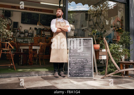 Full length shoot of a young waiter standing in door of a cafe. Young man with beard wearing an apron  standing with her arms cr Stock Photo