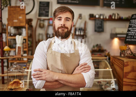 Portrait of young man wearing apron standing with his arms crossed in a coffee shop. Caucasian man with beard standing in a cafe Stock Photo