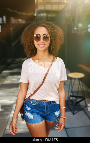 Stylish african woman wearing sunglasses posing outdoors. Beautiful young girl with curly hair in casuals walking on sidewalk. Stock Photo