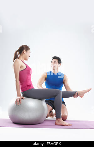 Woman leaning back on gym ball and lifting one leg held by man coaching her Stock Photo