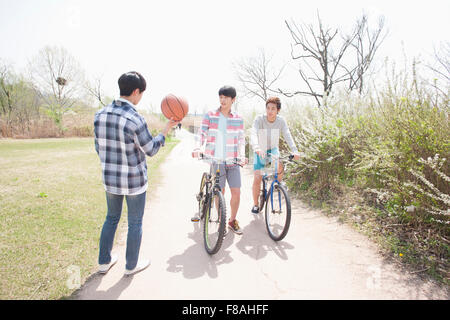 Back appearance of a man holding a basketball and two other men with their bikes on the road at the park Stock Photo