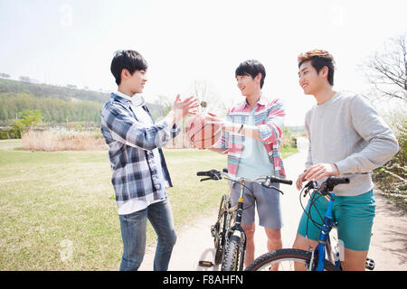 Man holding a basketball and two other men with their bikes on the road at the park Stock Photo