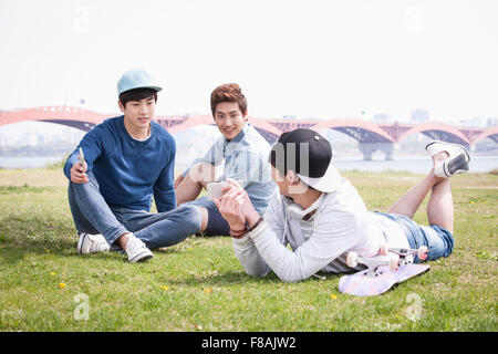 Two young men sitting on the grass and the other young man lying on his stomach on the grass at the park Stock Photo