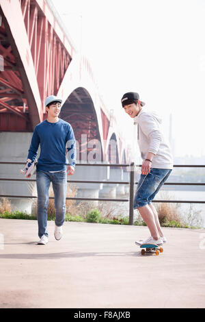Young man in a black cap skateboarding and the other man in a blue cap holding his skateboard under his arm and walking Stock Photo
