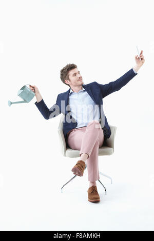 Businessman sitting on chair with his legs crossed, posing for sel-fie with watering can Stock Photo