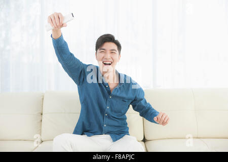 Smiling excited young man cheering holding hand with remote control sitting on sofa Stock Photo