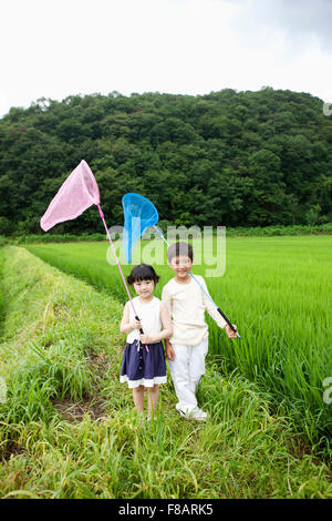 Smiling boy and girl standing with nets staring at front on lane through rice fields in spring Stock Photo
