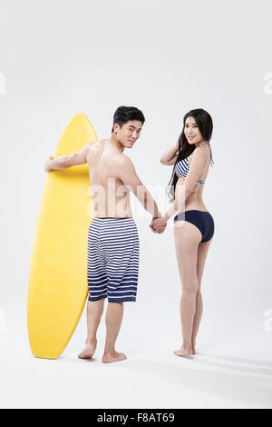 Backs of couple in swimsuits standing hand in hand with a surfing board looking back Stock Photo