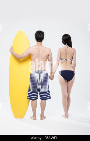 Backs of couple in swimsuits standing hand in hand with a surfing board Stock Photo