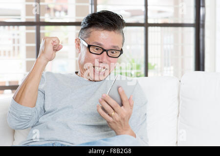 Portrait of excited 50s mature Asian man celebrating success while using smart phone, sitting on sofa at home. Stock Photo