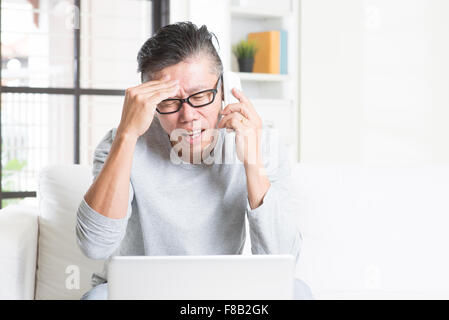 Portrait of 50s mature Asian man having problems and calling on phone, using computer internet working from home, sitting on sof Stock Photo