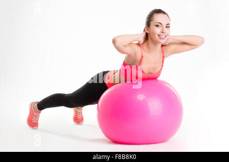 Pretty smiling young sportswoman in pink top and black leggings exercising with fitball isolated over white background Stock Photo