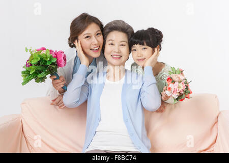 Grandmother seated on couch touching faces of mother and granddaughter behind the couch both holding flowers Stock Photo