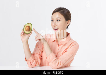 Adult woman in orange shirt seated at table holding half of avocado pointing with her finger Stock Photo