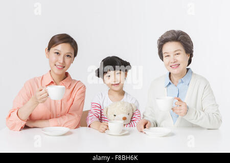 Young girl holding teddy bear seated at table between her mother and grandmother all holding a cup of tea each Stock Photo