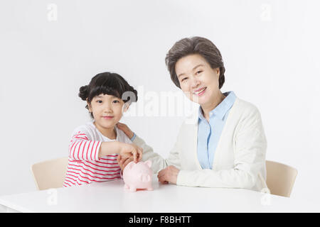 Young girl seated at table putting coins in the piggy bank and her grandmother seated right next to her both staring forward Stock Photo