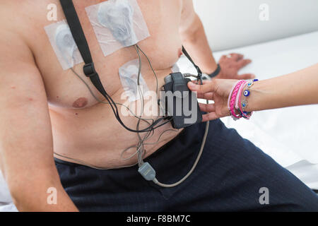Placing electrodes on the chest of a male patient, connected to a Holter monitor at her waist. A Holter monitor is a portable electrocardiograph (ECG) device. It can measure and record the electrical activity of a patient's heart over 24 hours, and detect Stock Photo