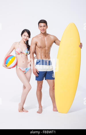 Woman in colorful bikini holding a beach ball under her arm and standing close to a muscular man holding a surfing board next Stock Photo