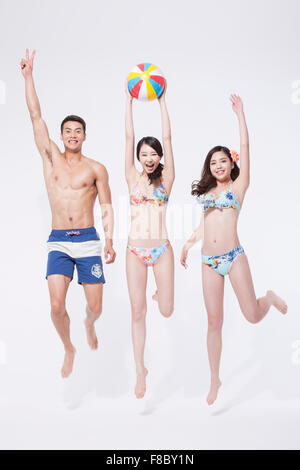 Two women and a men in beach wear jumping together with a beach ball in the woman in the middle of them Stock Photo
